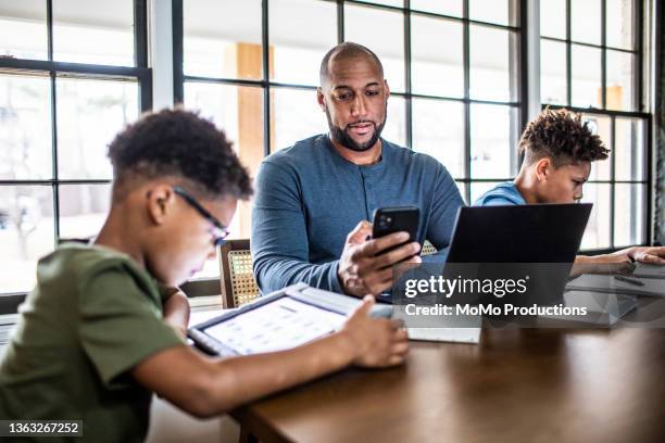 father working from home in residential kitchen with family in background - african ethnicity stock photos et images de collection