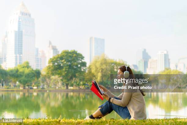 asian woman is happy relaxing by listen music and reading book at a city park in the morning, people relaxing in urban nature environments."r - mujer leyendo libro en el parque fotografías e imágenes de stock