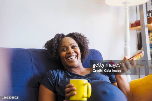 laughing pregnant woman with tea and smart phone on sofa - pregnant home stock pictures, royalty-free photos & images