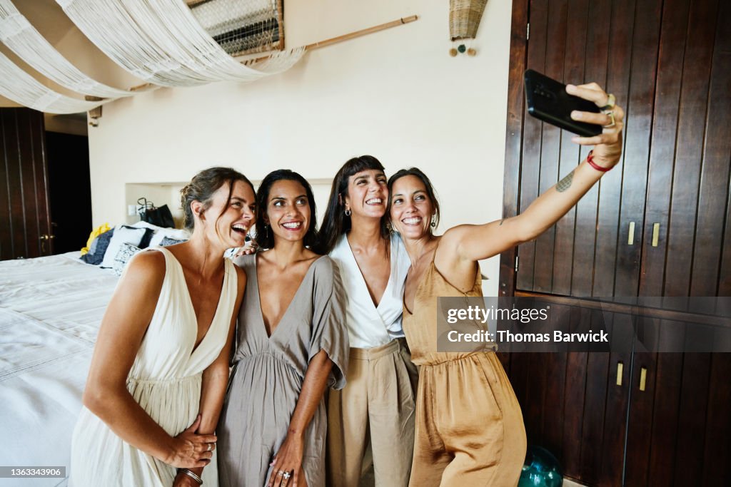 Medium wide shot of smiling and laughing bridesmaids taking selfie in luxury hotel suite before wedding