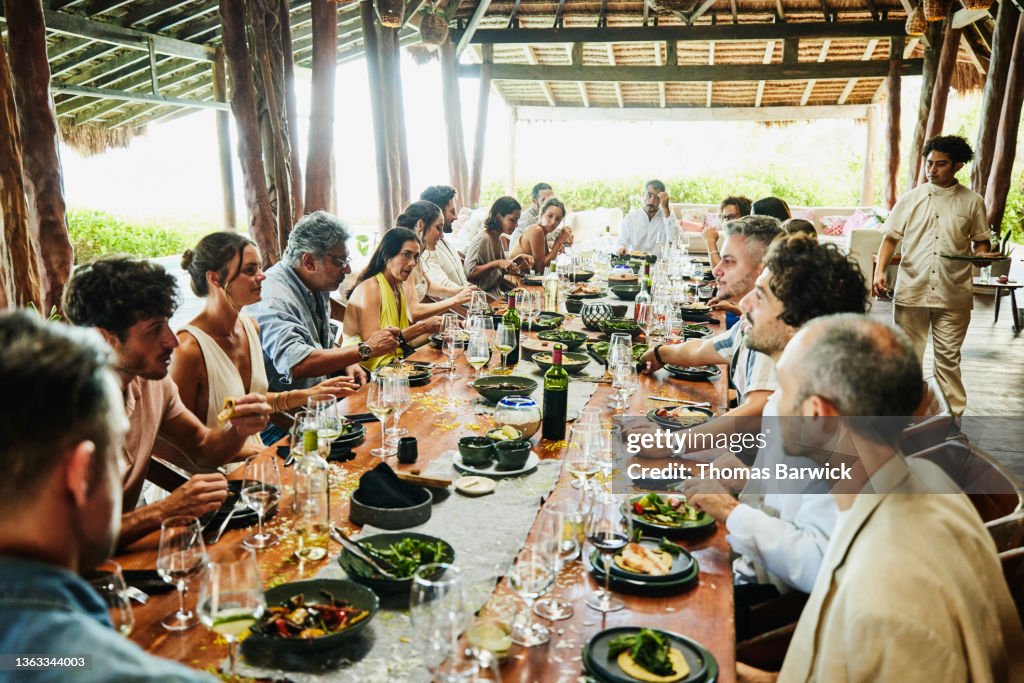 Wide shot of friends and family dining at long table at luxury tropical villa during wedding reception