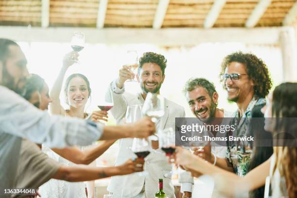 medium wide shot of smiling bride and groom toasting with wine glasses with guests during wedding reception dinner at tropical resort - medium group of people foto e immagini stock