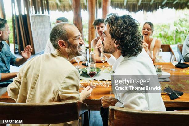 medium wide shot of gay couple eating piece of spaghetti together during wedding reception dinner at luxury tropical resort - medium group of people foto e immagini stock