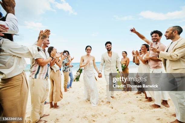 wide shot of bride and groom walking down aisle after wedding ceremony on tropical beach while friends and family celebrate - bride walking stock pictures, royalty-free photos & images