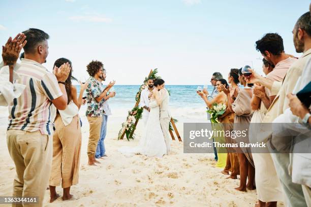 wide shot of bride and groom embracing while family and friends celebrate after wedding ceremony on tropical beach - wedding ceremony guests stock pictures, royalty-free photos & images