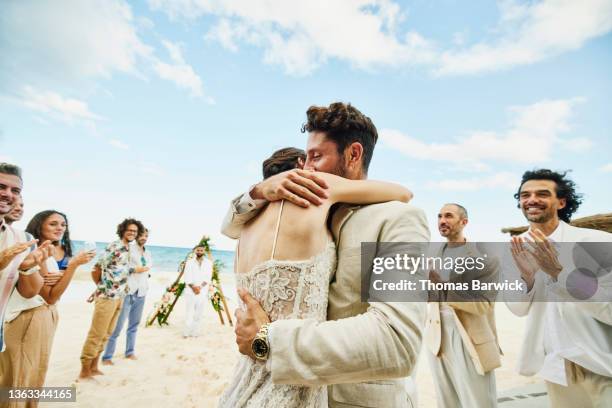 medium wide shot of bride and groom embracing in front of friends and family celebrating after wedding ceremony on tropical beach - trauung stock-fotos und bilder
