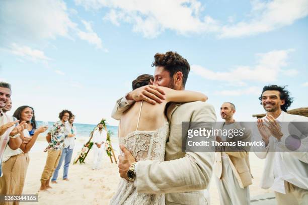 medium wide shot of bride and groom embracing in front of friends and family celebrating after wedding ceremony on tropical beach - matrimonio - fotografias e filmes do acervo