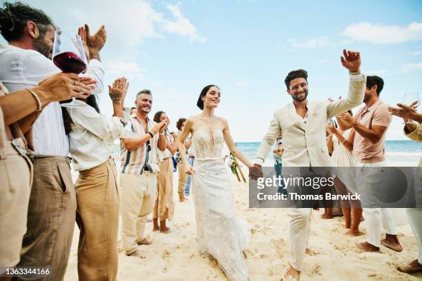 wide shot of bride and groom walking down aisle after wedding ceremony on tropical beach while friends and family celebrate - koninklijke bruiloft stockfoto's en -beelden