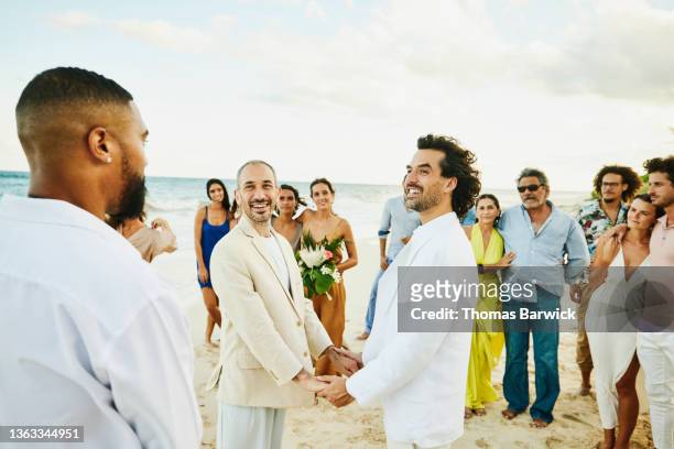 medium wide shot of smiling gay couple holding hands while getting married in front of friends and family on tropical beach - medium group of people foto e immagini stock