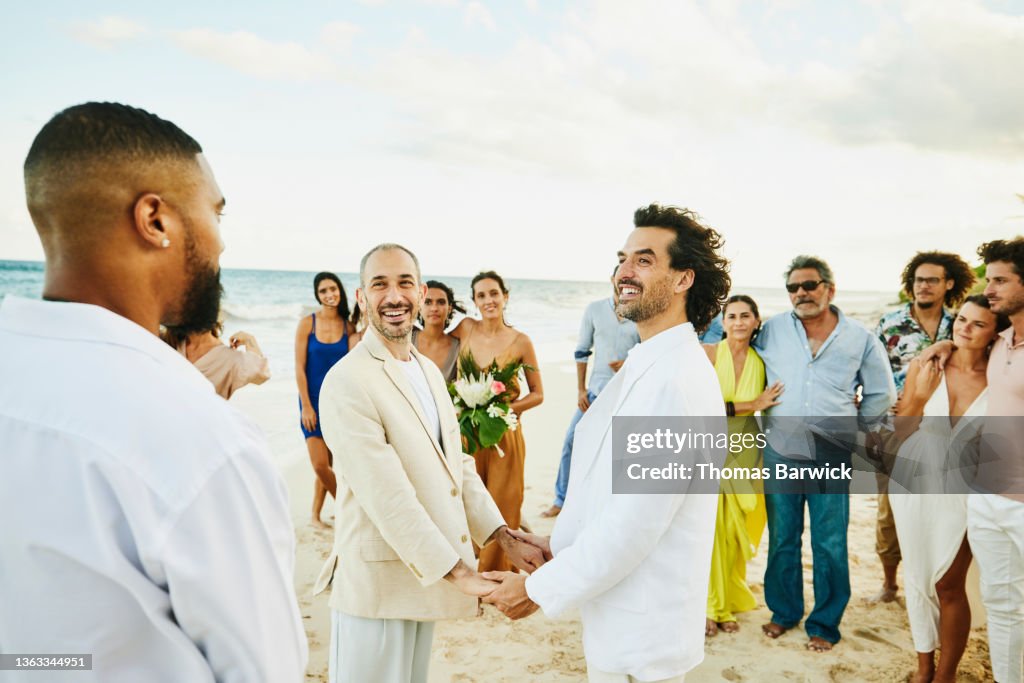Medium wide shot of smiling gay couple holding hands while getting married in front of friends and family on tropical beach