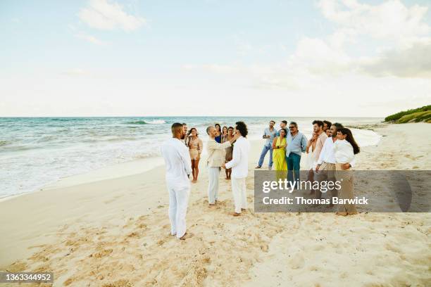 wide shot of smiling gay couple getting married in front of friends and family on tropical beach - homohuwelijk stockfoto's en -beelden