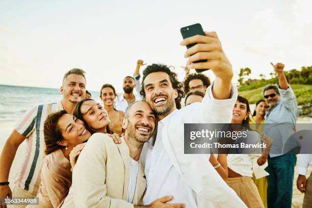 medium wide shot of smiling gay couple taking selfie with friends and family after wedding ceremony on tropical beach - wedding people stock pictures, royalty-free photos & images