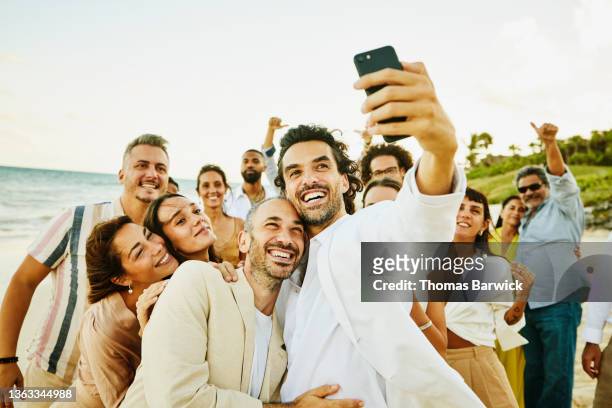 medium wide shot of smiling gay couple taking selfie with friends and family after wedding ceremony on tropical beach - medium group of people imagens e fotografias de stock