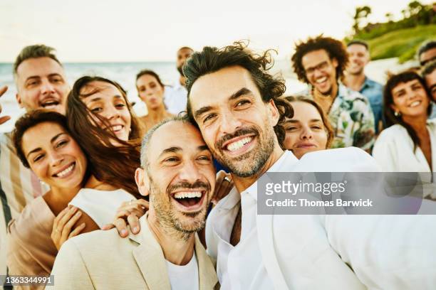 medium shot of smiling gay couple taking selfie with friends and family after wedding ceremony on tropical beach - medium group of people imagens e fotografias de stock
