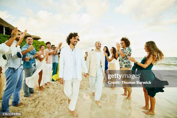 wide shot of gay couple walking down aisle after wedding ceremony on tropical beach while friends and family celebrate - civil partnership stock pictures, royalty-free photos & images