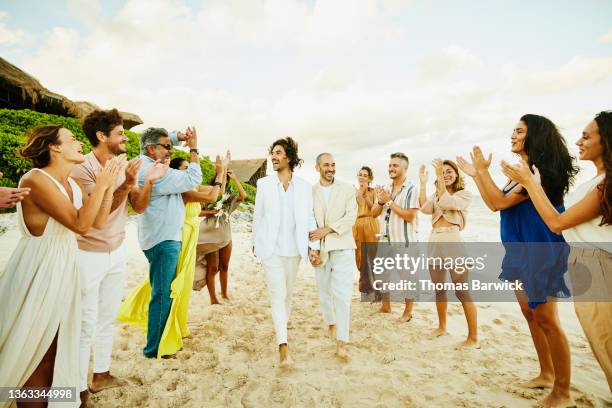 wide shot of gay couple walking down aisle after wedding ceremony on tropical beach while friends and family celebrate - homohuwelijk stockfoto's en -beelden