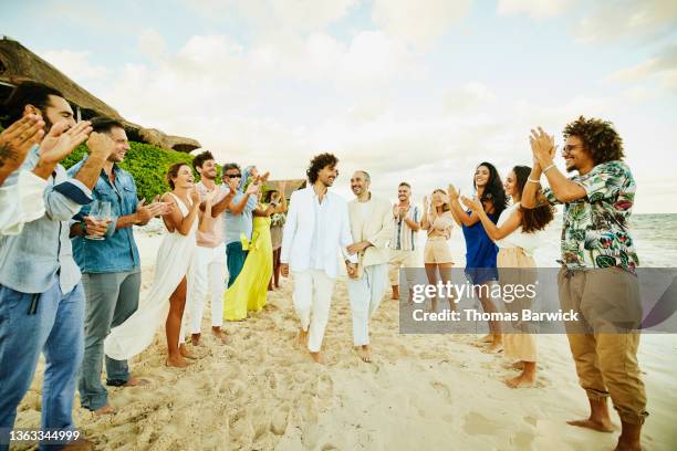 wide shot of gay couple walking down aisle after wedding ceremony on tropical beach while friends and family celebrate - destination wedding imagens e fotografias de stock