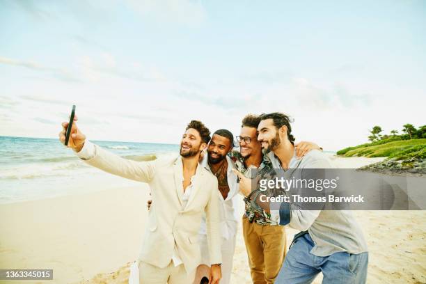medium wide shot of smiling friends taking selfie with smart phone after tropical beach wedding - medium group of people foto e immagini stock