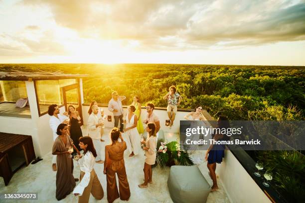 wide shot elevated view of smiling and laughing friends and family celebrating on rooftop deck at sunset after wedding at tropical resort - privilegien stil stock-fotos und bilder