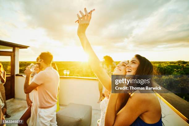 medium shot of smiling bride taking selfie with friend during rooftop party at sunset after wedding at tropical resort - sunlight through drink glass stock-fotos und bilder