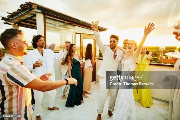 Wide shot of smiling bride and groom celebrating with friends on rooftop deck at sunset after wedding at tropical resort