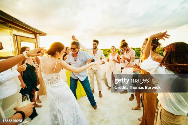 wide shot of smiling bride dancing with father and celebrating with friends on rooftop deck at sunset after wedding at tropical resort - banquete de boda fotografías e imágenes de stock