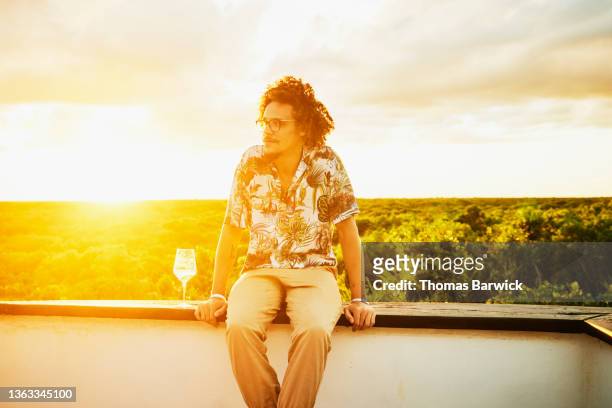 Medium wide shot of man sitting on edge of rooftop deck at sunset at tropical resort