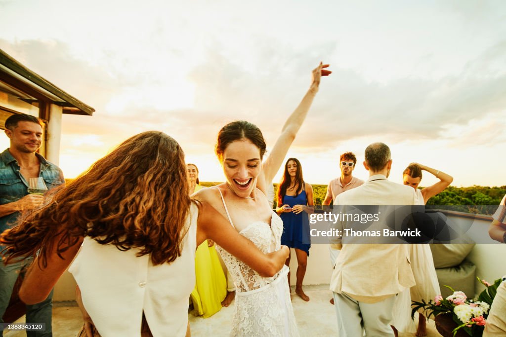 Medium wide shot of smiling bride and friend dancing during party on rooftop deck after wedding at tropical resort