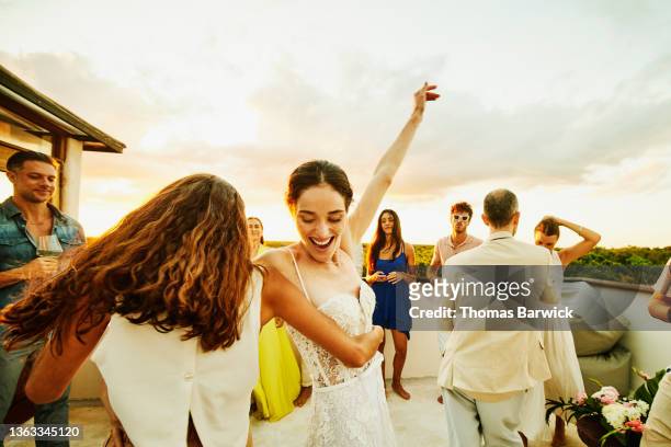 Medium wide shot of smiling bride and friend dancing during party on rooftop deck after wedding at tropical resort