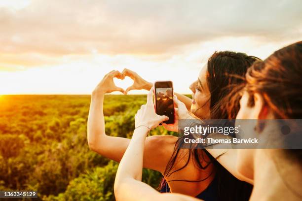 medium shot of woman taking photo of smiling friend making heart shape with hands over sunset during rooftop party - premium access image only stock-fotos und bilder