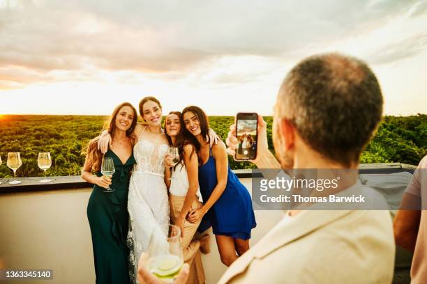 medium wide shot of man taking photo of bride and friends during rooftop party after wedding at tropical resort - hochzeitsgesellschaft stock-fotos und bilder