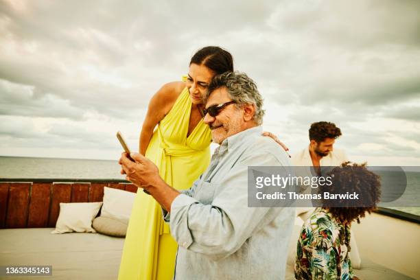 medium wide shot of senior couple looking at photo on smart phone during sunset rooftop party at tropical resort - premium acess stock-fotos und bilder