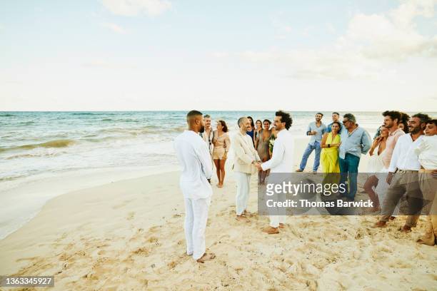 wide shot of smiling gay couple holding hands while getting married in front of friends and family on tropical beach - civil partnership stock pictures, royalty-free photos & images