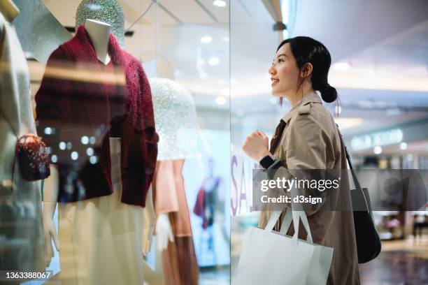 beautiful smiling young asian woman carrying a shopping bag, standing outside a boutique looking at window display in a shopping mall in city. sale season, festive shopping atmosphere - erotique chic photos et images de collection