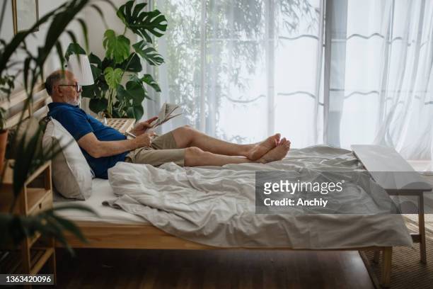 elderly man reading a newspaper during a relaxing day at home - read and newspaper and bed stock pictures, royalty-free photos & images