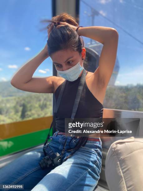 young peruvian woman wears a blue surgical face mask and holds her brown hair with her hands inside a cable car cabin in medellin, colombia - altitude sickness stock pictures, royalty-free photos & images