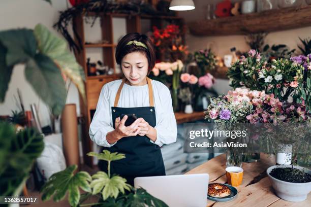 asian female florist, owner of small business flower shop, using smartphone while working on laptop against flowers and plants. checking stocks, taking customers orders, selling products online. daily routine of running a small business with technology - asia lady selling flower stock pictures, royalty-free photos & images