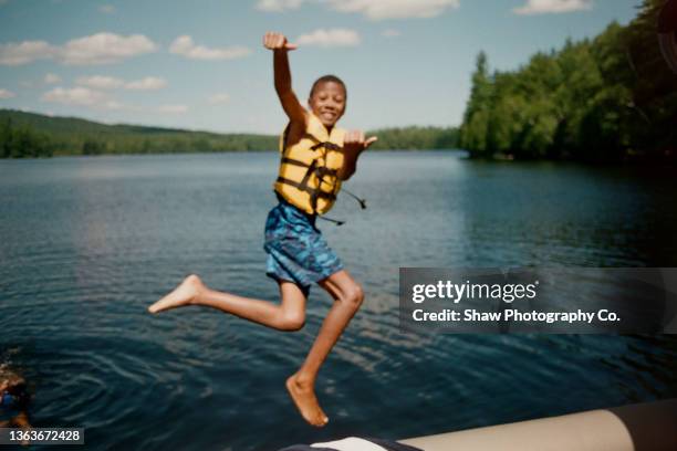 an out of focus photo of an african-american boy jumping off a cliff into a lake with a yellow life jacket on. his arms are up and he is smiling - camping kids photos et images de collection