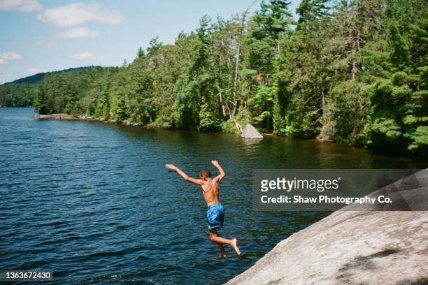 a portrait of an african-american boy jumping off a rock into a blue lake in the adirondacks with trees surrounding the blue lake - adirondack state park stock-fotos und bilder