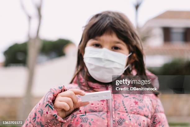 little girl in the street, wearing unfocused white mask, holding a negative antigen test. concept covid-19, ihu, delta, omicron, detection test and pandemic. - small cotton plant stockfoto's en -beelden