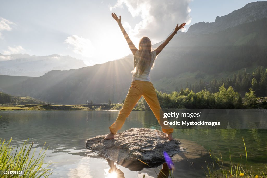 Young woman arms up on rock above alpine lake at sunrise, happy morning concept