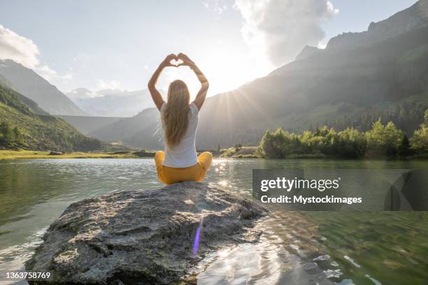 young woman sits on rock above lake and makes a heart with hands - friedenssymbol bildbanksfoton och bilder