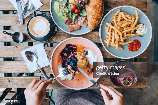 overhead view of young asian woman sitting in cafe, eating pancakes with blueberries and whipped cream, with english breakfast and french fries served by the side on the dinning table. people and food concept, eating out lifestyle - fine dining table side view stock-fotos und bilder