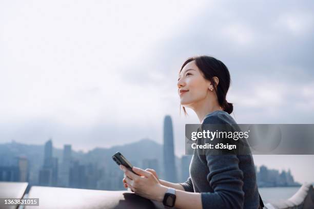 beautiful young asian woman using smartphone by the promenade in the city, looking ahead with smile, with the view of urban city skyline. optimistic and hopeful emotion. lifestyle and technology - happy smile with phone foto e immagini stock