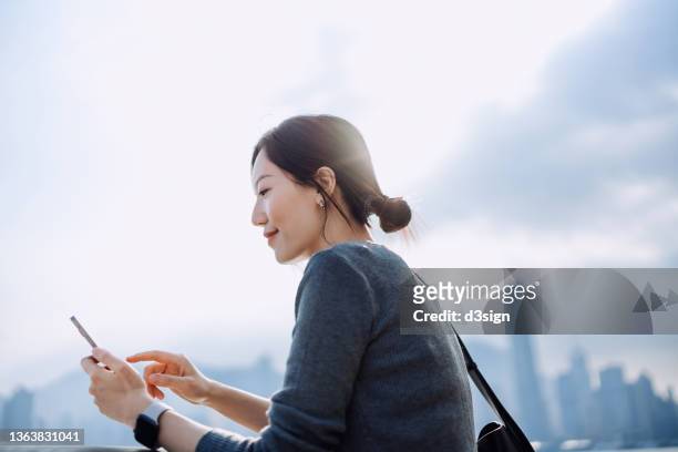 smiling young asian businesswoman standing by the promenade, with the view of urban city skyline, managing finance and investment with online banking on smartphone. smart banking anytime anywhere with technology concept - asia skyline stock pictures, royalty-free photos & images