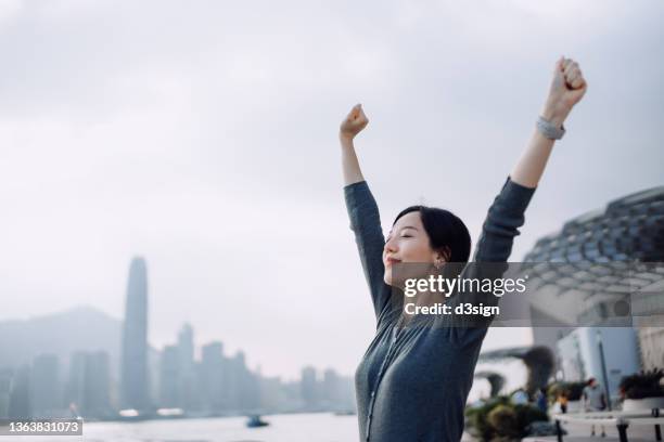 beautiful young asian woman with her eyes closed, taking a deep breath and stretching arms against victoria harbour and urban city skyline - erleichterung stock-fotos und bilder