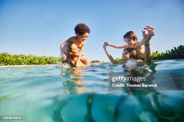 wide shot of smiling sons riding on mothers backs while playing in pool at tropical resort - family pool stock-fotos und bilder