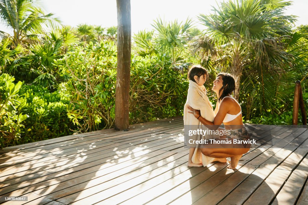 Wide shot of mother drying of son with towel on deck of luxury suite at tropical resort