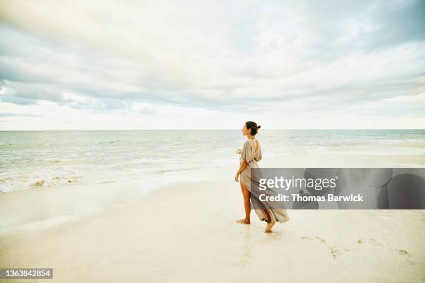 wide shot of woman walking on beach and enjoying sunset at tropical resort - beige dress stock pictures, royalty-free photos & images