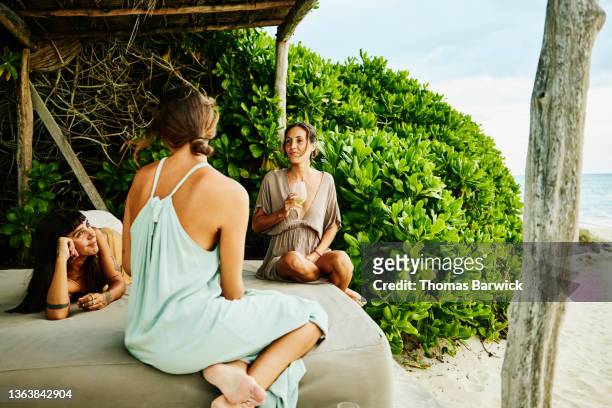 medium wide shot of female friends in discussion while relaxing in cabana on beach at tropical resort - medium group of people foto e immagini stock